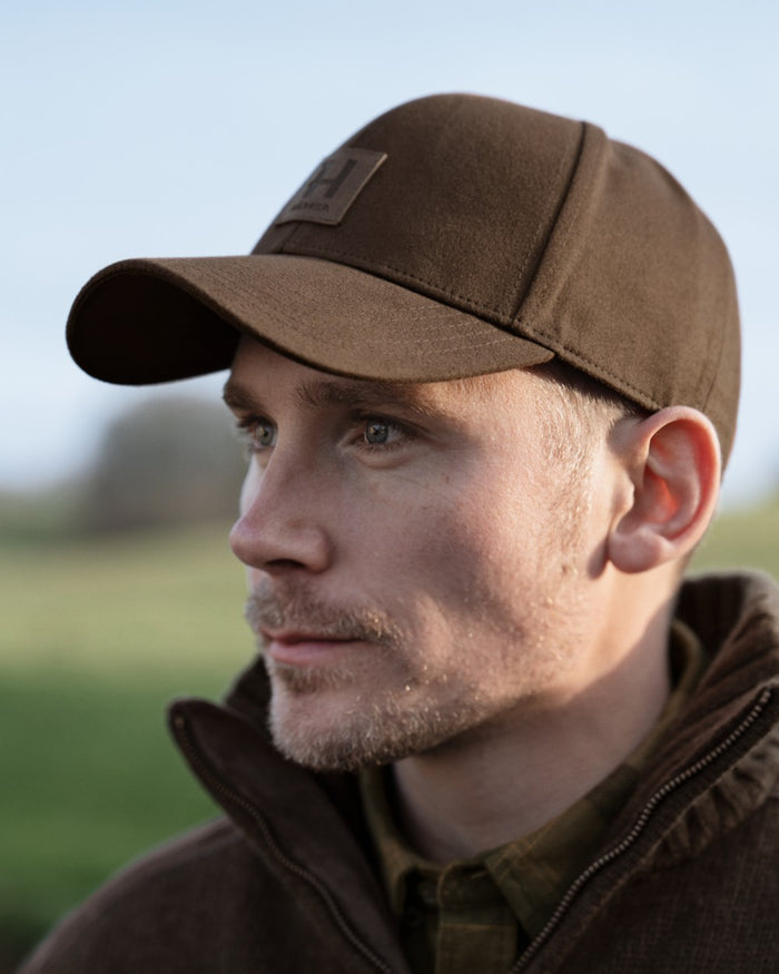 A man in profile wearing a brown baseball cap with a small rectangular logo patch on the front, photographed outdoors against a softly blurred green and blue background. #colour_dark-brown