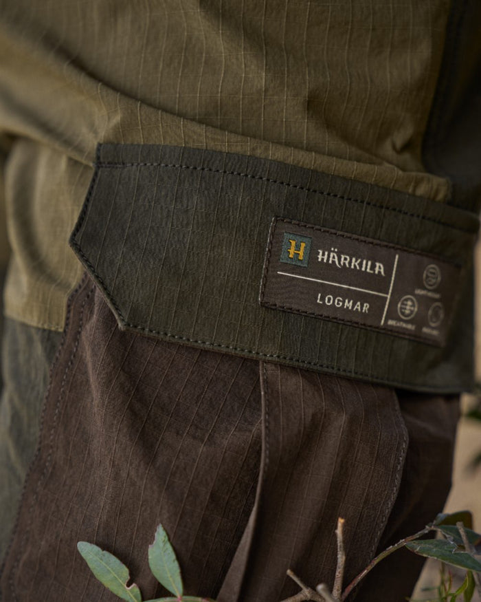 Dark Olive Willow Green Coloured Harkila Logmar Lightweight Zip Off Trousers on forest background #colour_dark-olive-willow-green
