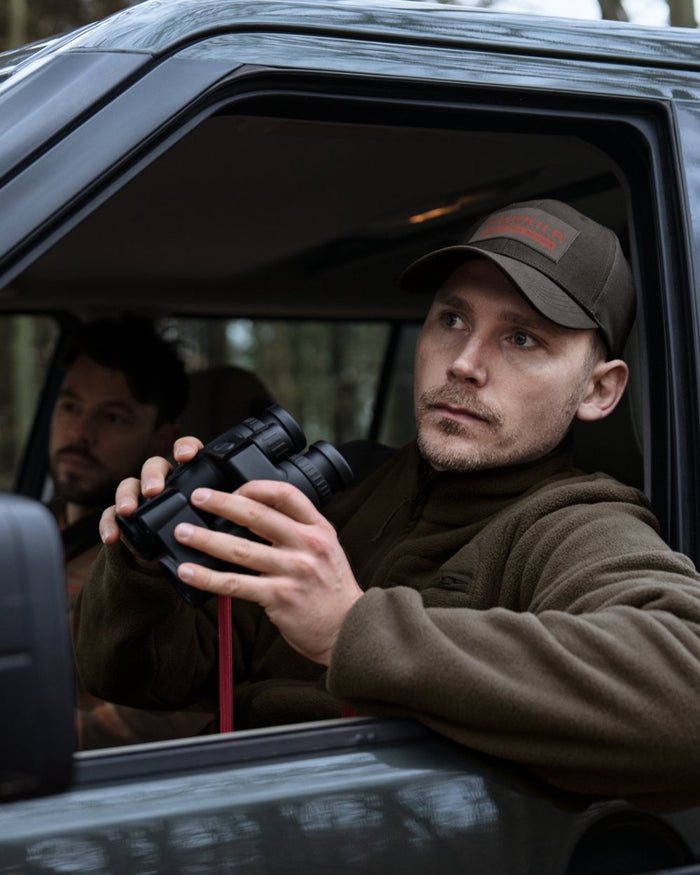 A man sitting in the driver's seat of a dark vehicle looks out the window while holding black binoculars. He is wearing a brown fleece jacket and a brown cap with a red logo. #colour_shadow-brown