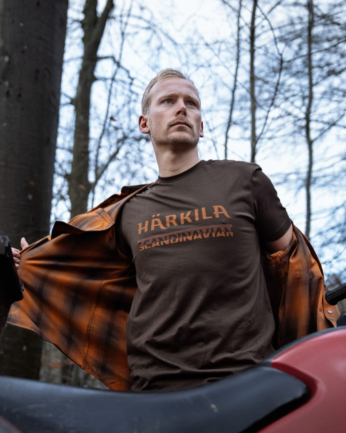 A man wearing a brown t-shirt with printed in orange-brown, holding open an orange-brown plaid flannel shirt in an outdoor forest setting. #colour_shadow-brown