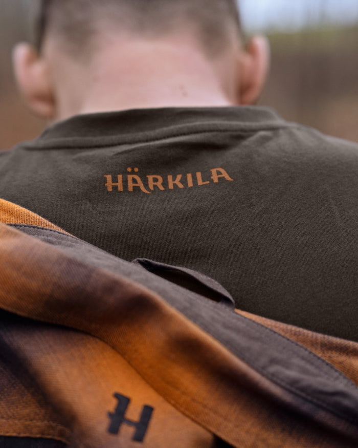 Close-up back view of a person wearing a dark brown Härkila shirt, with a brown and orange plaid scarf draped over the shoulders and the Härkila logo embroidered on the upper back. #colour_shadow-brown