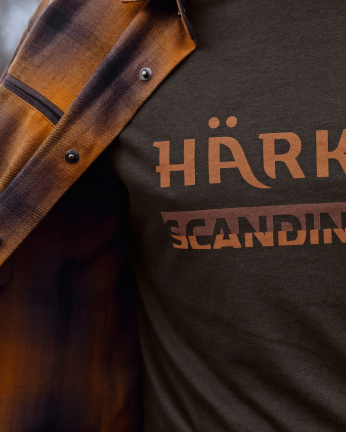 Close-up of a person wearing a dark brown heather t-shirt with a copper-brown graphic print that reads, with an open brown and black plaid flannel shirt draped over the shoulders. #colour_shadow-brown