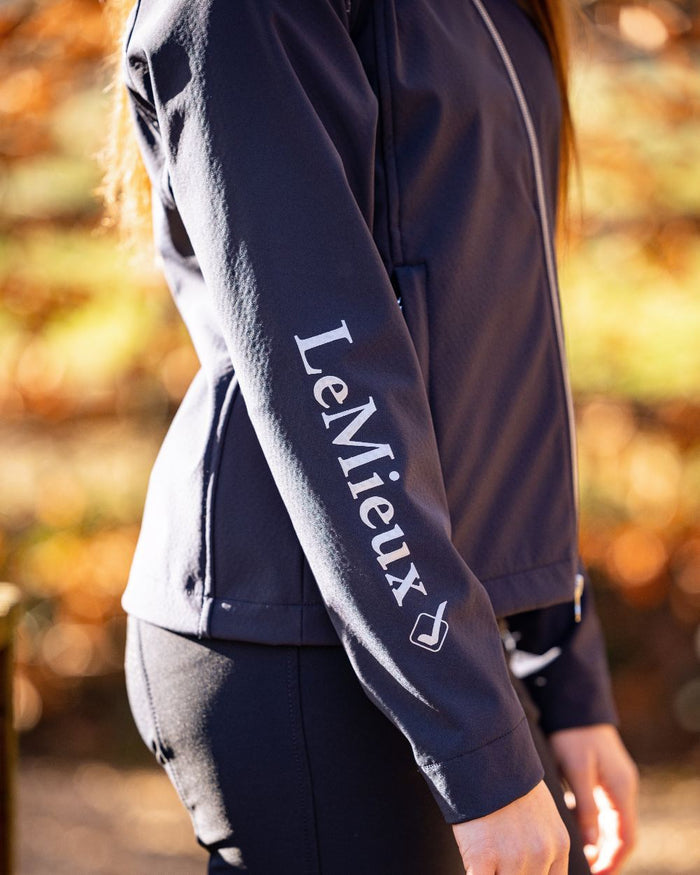 Navy coloured LeMieux Young Rider Elite Soft Shell Jacket with trees in background #colour_navy