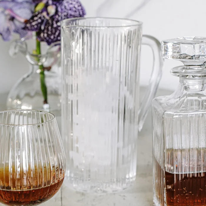 The Vietri Natalia Pitcher filled with ice, accompanied by a matching glass containing a brown liquid and a faceted decanter holding amber liquid, are arranged on a table. In the background, flowers in a vase appear softly blurred.