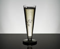 A Tom Dixon Puck Flute Glass from the Set of 2, filled with sparkling wine and rising bubbles, stands on a reflective black surface against a minimalist gray background.