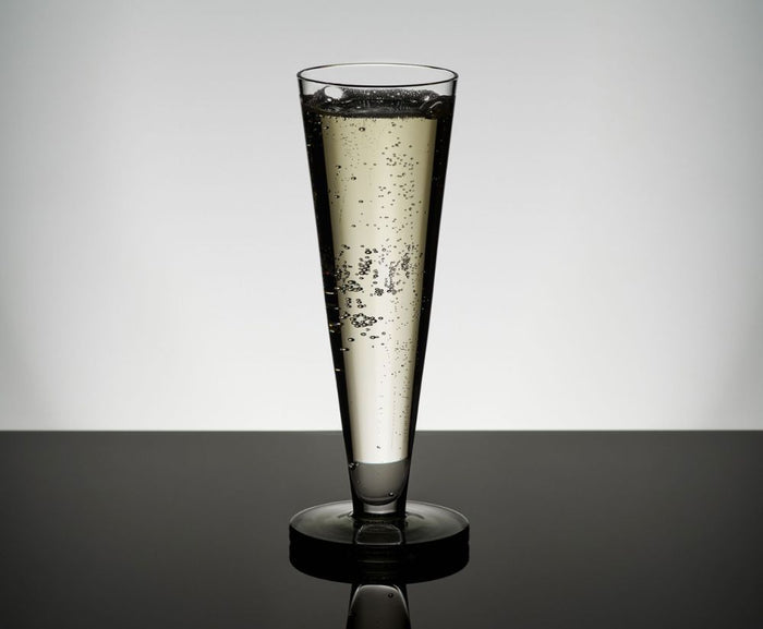 A Tom Dixon Puck Flute Glass from the Set of 2, filled with sparkling wine and rising bubbles, stands on a reflective black surface against a minimalist gray background.
